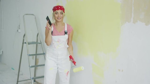 Side view of young blond female in white overalls and red bandana painting plastered wall pastel yellow with roller and talking on mobile phone in hand.