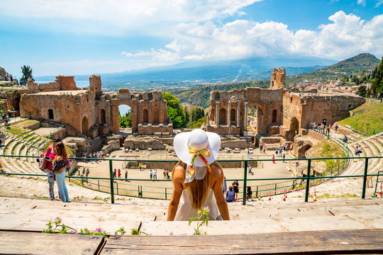 Jule 04, 2018. Taormina, Italy. Beautiful girl in a long white dress and white summer hat sitting at the caldron of the ancient Greek Theater of Taormina in Italy.
