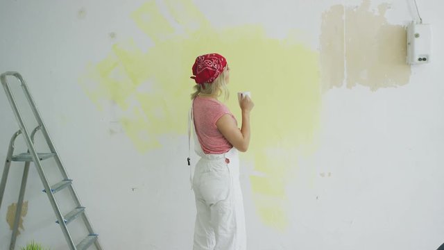 Back view of young blond woman in white overalls standing relaxed with cup and inspecting results of work on half-painted pastel yellow wall