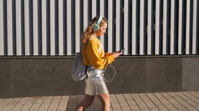 Portrait of young cute attractive young girl in urban background listening to music with headphones. Woman wearing yellow blouse and silver skirt