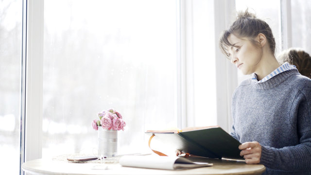 A young brunette girl in a cafe sitting near the window dressed in a blue pullover is holding a black notebook thoughtfull look