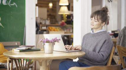 Fototapeta na wymiar A young smiling cute happy brunette girl dressed in a grey pullover is working with a laptop in a cafe