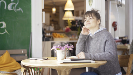 A young smiling cute brunette girl dressed in a grey pullover is sitting in a cafe thinking over