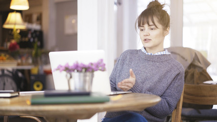 A young smiling delightful cute happy brunette girl dressed in a grey pullover is working with a laptop in a cafe