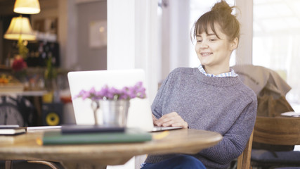 Obraz na płótnie Canvas A young smiling delightful cute happy brunette girl dressed in a grey pullover is working with a laptop in a cafe