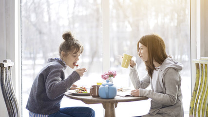 Two young adult female friends eating salad drinking tea sitting near a big window indoors in a cafe