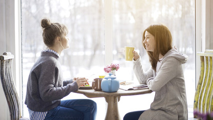Two young adult female friends eating salad drinking tea sitting near a big window indoors in a cafe