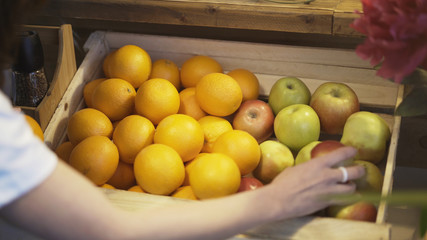 Closeup of a hand of brunette girl no face is taking an apple in her hand from a wooden box full of oranges and apples in a market place.