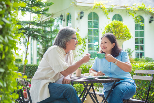 Retired Asian Couple Sitting At Table In Garden And Drinking Coffee