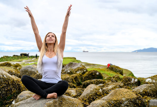 Young woman sitting on rock and her arms are raised into the air at the intertidal zone of Vancouver, British Columbia