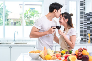 Obraz na płótnie Canvas Asian lovers or couples kissing forehead and drinking wine in kitchen room at home. Love and happiness concept Sweet honeymoon and Valentine day theme