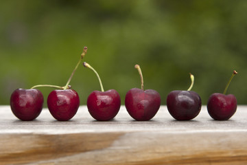 Six ripe red cherries with stems lined up in a row after harvest. Sweet, dark cherry fruits on wood outside.
