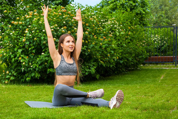 A dark-haired woman coach in a sporty short top and gym leggings does stretching the legs on the yoga mat, hands are raised upwards,  on a summer day in a park on a green lawn