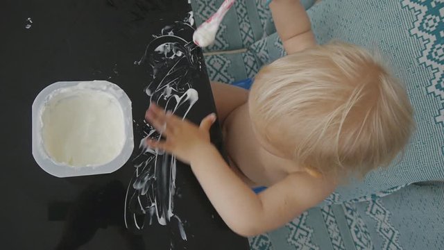 Baby eating yogurt making a mess. Blond naked baby girl eats yogurt with a spoon smearing it on the black glass table sitting on the plastic potty
