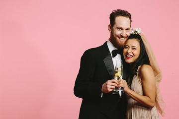 A smiling portrait of a bride and groom hugging and cheering with champagne glasses, isolated on pink