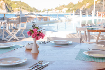 Fototapeta na wymiar Summer terrace of the restaurant. On the tables laying with white plates of wine glasses and white vases with pink tropical flowers.flowers on an empty outdoor restaurant table at a seaside resort in