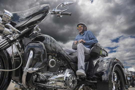 Elderly man sits on the back of a fancy three-wheeled motorcycle on a dark, stormy day 