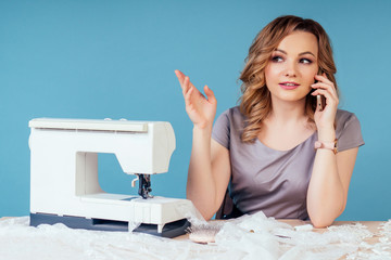 attractive blonde hairstyle woman seamstress tailor ( dressmaker) talking on the phone with a client behind the table with sewing machine on a blue background in the studio