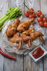 Baked chicken legs and wings lie on the table. Nearby are cherry tomatoes, celery, parsley and spices.