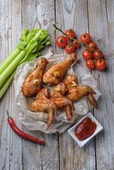Baked chicken legs and wings lie on the table. Nearby are cherry tomatoes, celery, parsley and spices.