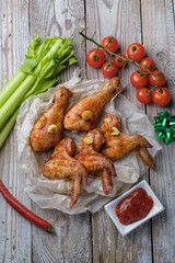 Baked chicken legs and wings lie on the table. Nearby are cherry tomatoes, celery, parsley and spices.