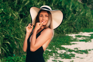 Portrait of a cute young attractive girl on the beach wearing a swimsuit, wearing a fashionable hat, covering it from the sun, playing her lips playfully, smiling and enjoying the holiday in the