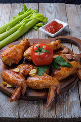 Baked chicken legs and wings lie on the table. Nearby are cherry tomatoes, celery, parsley and spices.