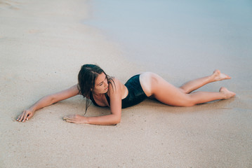 Fototapeta premium A sexy young girl with a smooth even skin in a fashionable black swimsuit lies on wet sand. The concept of leisure, travel, people.