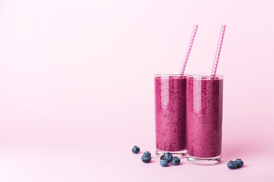 Two glasses of blueberries smoothie with straws on pink background. Healthy summer drink.