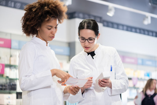 Two female pharmacists reading the information from the pack of a new product while comparing two different medicines regarding indications and side effects 