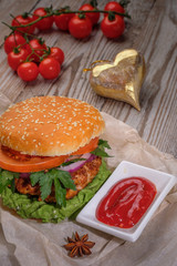The hamburger lies on an old wooden table. On the table is a hamburger, tomato sauce, cherry tomatoes, a jar of spices.