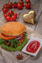 The hamburger lies on an old wooden table. On the table is a hamburger, tomato sauce, cherry tomatoes, a jar of spices.