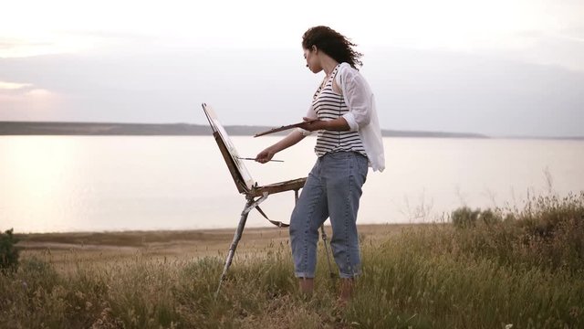Full length footage of a beautiful girl in white shirt and jeans working outdoors. Paints using an easel and oil paints on the meadow in front the lake. Wind blowing, morning