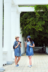 Fototapeta premium Two young beautiful girls are walking along a green park with white columns. Day, the sun. Travelers, tourists, laugh and take pictures on a smartphone and camera.