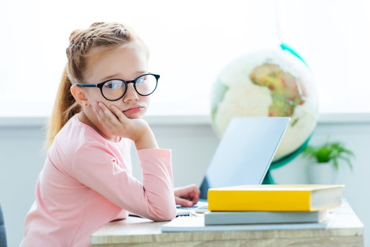 bored child in eyeglasses looking at camera while studying with laptop and books