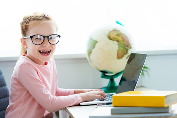adorable happy schoolchild in eyeglasses studying with laptop and books at home