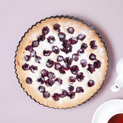 Whole homemade cherry cake on pastel pink background with teapot and cup of tea.