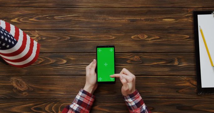 View from the top on the woman's hands scrolling and taping on the black smartphone with green screen on the wooden desk with American flag and blank paper sheet with a pencil. Vertically. Chroma key
