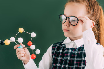 beautiful little schoolgirl in eyeglasses holding molecular model while standing near chalkboard