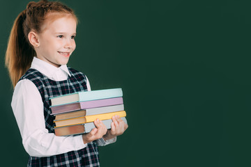 beautiful smiling little schoolgirl holding pile of books and looking away while standing near blackboard