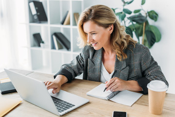 attractive businesswoman using laptop and writing something to notebook in office