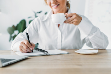 cropped image businesswoman holding cup of coffee and writing to notebook in office
