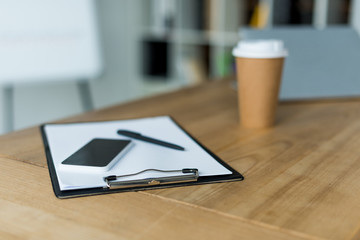 smartphone and disposable coffee cup with clipboard on wooden table in office