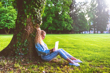 young blonde woman sitting ont he grass using notebook leaning against a trunk in a city park -...