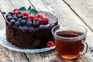 Homemade chocolate cake decorated with black and red raspberries on glass plate with cup of tea
