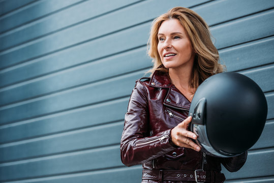 attractive woman in leather jacket holding motorcycle helmet on street and looking away