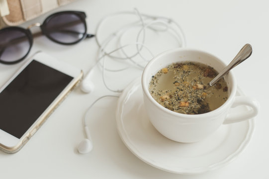 Cup of herbal tea with camomile, smartphone, earphones and sunglasses on white table. Tea break concept