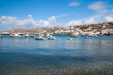 Fototapeta na wymiar Boats on sea water in Mykonos, Greece. Sea village on cloudy blue sky. White houses on mountain landscape with nice architecture. Summer vacation on mediterranean island. Wanderlust and travelling