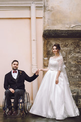 Groom on the wheelchair holds bride's hand standing before old house on the street