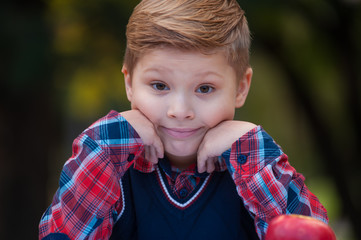 Close-up portrait of a charming little brown-eyed boy first-grader in a shirt and waistcoat looking at the camera on a background of blurred greens in the park.